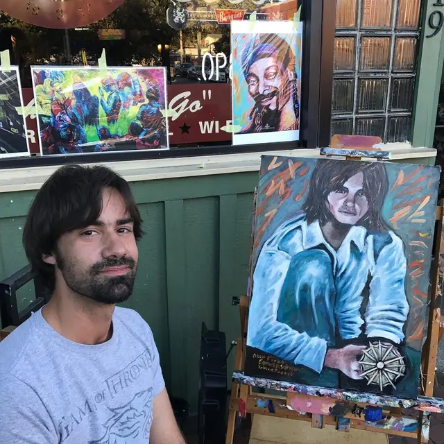 Toby seated on the sidewalk beside an easel holding one of his portraits